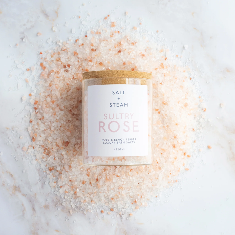 The image shows a jar of Salt + Steam Sultry Rose Bath Salts, elegantly styled with a cork lid. The jar rests on a bed of pink and white bath salts scattered across a smooth marble surface. The minimalist label highlights the product name and details, featuring "Rose & Black Pepper Luxury Bath Salts" with a net weight of 432g. This arrangement emphasizes a luxurious, natural, and soothing aesthetic, perfect for self-care and relaxation.