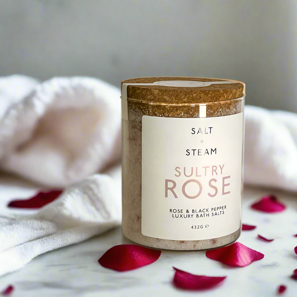 The image shows a jar of Salt + Steam Sultry Rose Bath Salts, elegantly styled with a cork lid. The jar rests on a bed of pink and white bath salts scattered across a smooth marble surface. The minimalist label highlights the product name and details, featuring "Rose & Black Pepper Luxury Bath Salts" with a net weight of 432g. This arrangement emphasizes a luxurious, natural, and soothing aesthetic, perfect for self-care and relaxation.