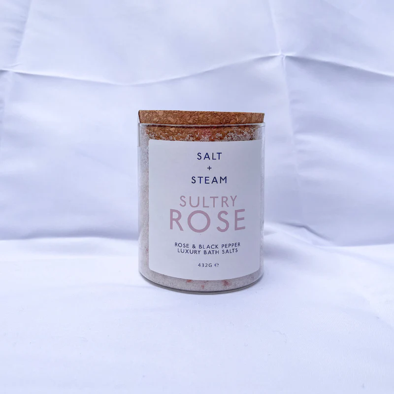 The image shows a jar of Salt + Steam Sultry Rose Bath Salts, elegantly styled with a cork lid. The jar rests on a bed of pink and white bath salts scattered across a smooth marble surface. The minimalist label highlights the product name and details, featuring "Rose & Black Pepper Luxury Bath Salts" with a net weight of 432g. This arrangement emphasizes a luxurious, natural, and soothing aesthetic, perfect for self-care and relaxation.