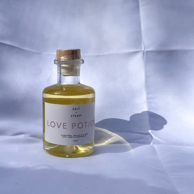 The image features a glass bottle labeled "Love Potion" by Salt + Steam, containing a vibrant golden yellow oil. The bottle has a wooden cork-style cap and a minimalist white label with soft pink text. It is set against a textured light backdrop with natural shadows, creating a clean and simple aesthetic. The soft lighting highlights the clarity and rich color of the oil, evoking a sense of warmth and luxury