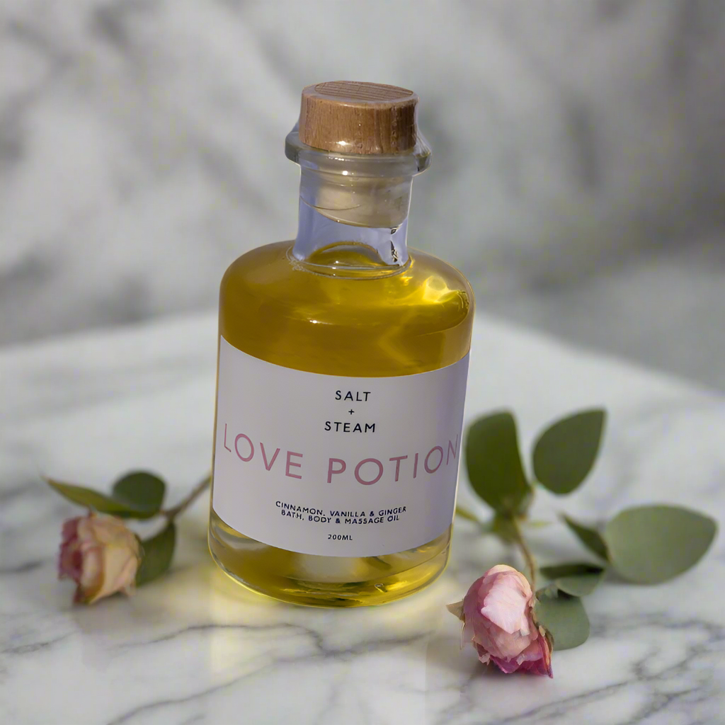 The image features a glass bottle labeled "Love Potion" by Salt + Steam, containing a vibrant golden yellow oil. The bottle has a wooden cork-style cap and a minimalist white label with soft pink text. It is set against a textured light backdrop with natural shadows, creating a clean and simple aesthetic. The soft lighting highlights the clarity and rich color of the oil, evoking a sense of warmth and luxury.