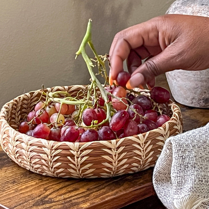 Handwoven pine needle basket filled with fresh red grapes, showcasing its natural, artisanal design. Perfect for serving fruit or adding rustic charm to your kitchen or dining decor.