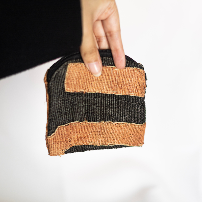 Handwoven natural fiber pouch with a zipper closure, featuring a bold striped design in black and earthy orange tones, held for scale to showcase its compact, artisanal craftsmanship.