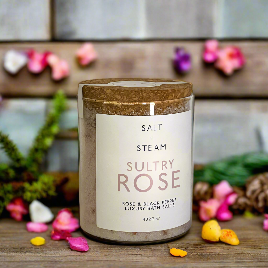 The image shows a jar of Salt + Steam Sultry Rose Bath Salts, elegantly styled with a cork lid. The jar rests on a bed of pink and white bath salts scattered across a smooth marble surface. The minimalist label highlights the product name and details, featuring "Rose & Black Pepper Luxury Bath Salts" with a net weight of 432g. This arrangement emphasizes a luxurious, natural, and soothing aesthetic, perfect for self-care and relaxation.