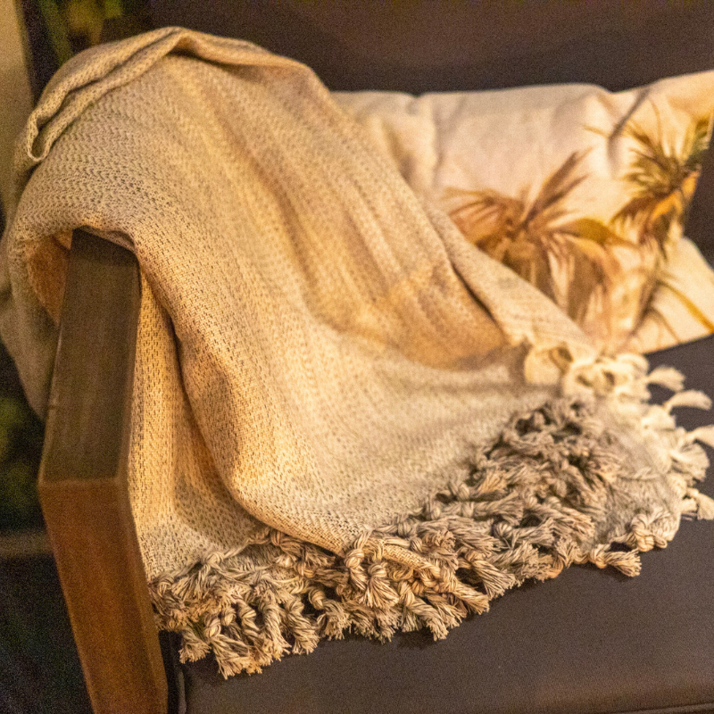 Cozy cream and gray throw blanket with tasseled fringe, draped over a wooden armchair in a warm and inviting setting, complemented by a decorative tropical pillow.
