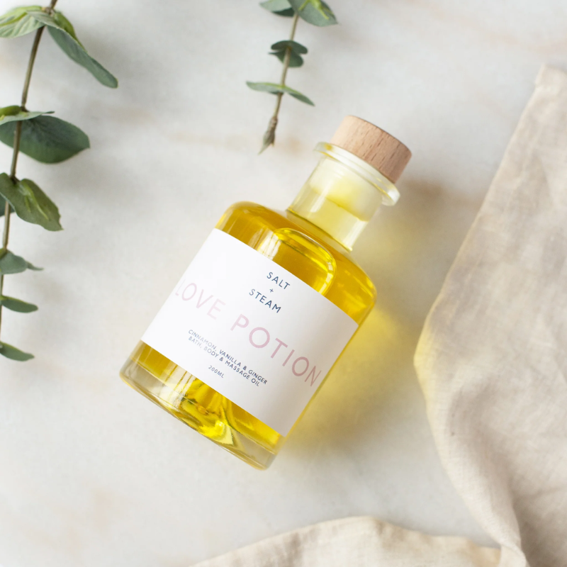 The image showcases a beautifully styled bottle of bath and body oil labeled "Love Potion" by Salt + Steam. The bottle is made of clear glass, revealing a rich golden yellow oil inside. It features a wooden cap and a minimalist white label with soft pink text, adding an elegant and luxurious touch. The bottle is placed on a light marble or stone surface, accented by sprigs of green foliage and a beige linen fabric. The overall aesthetic is clean, calming, and natural, evoking a sense of relaxation and indul