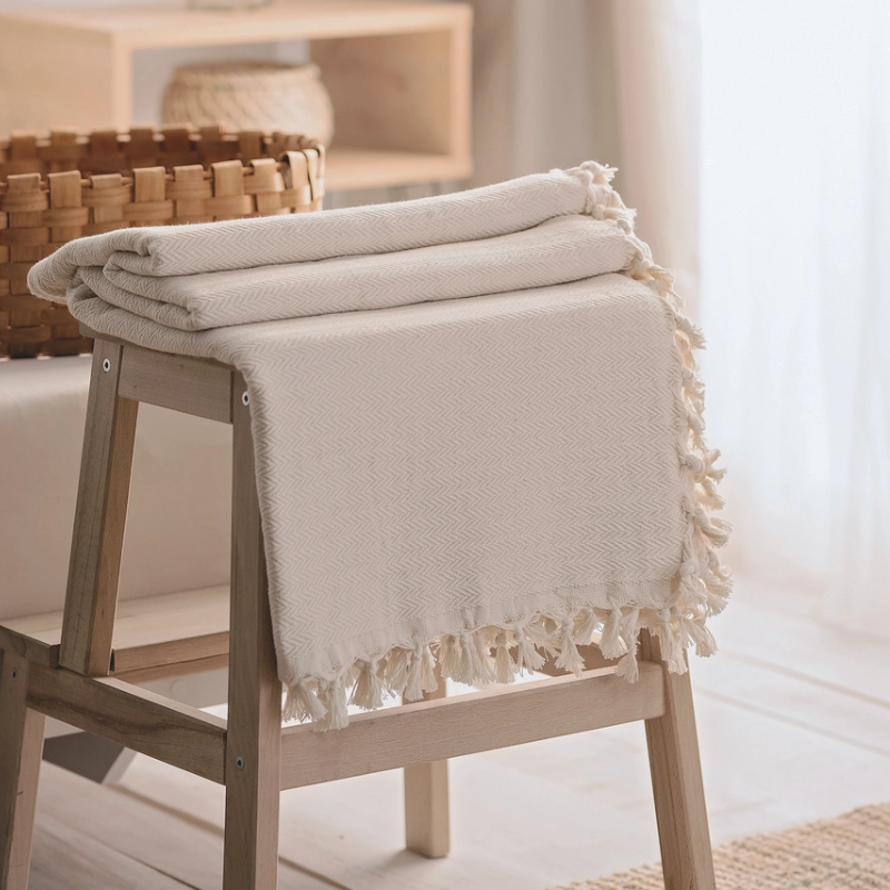 Folded cream farmhouse throw blanket with a chevron pattern and tassel fringe, draped over a wooden stool in a cozy, rustic setting.
