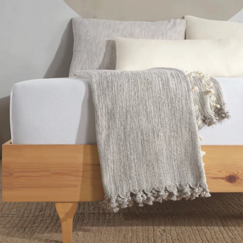 Gray farmhouse throw blanket with a textured pattern and tassel fringe, draped over a wooden bedframe in a cozy, minimalist bedroom setting.