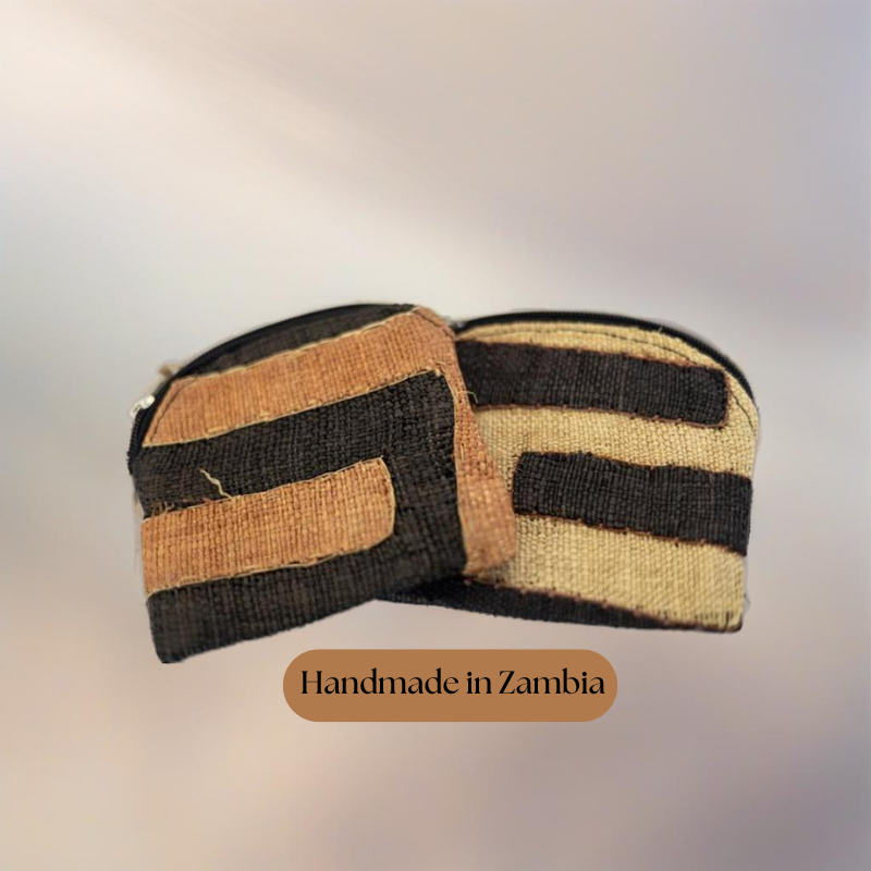 Two handwoven natural fiber pouches with black and earthy orange or beige stripes, displayed against a soft, blurred gradient background. Text overlay reads 'Handmade in Zambia,' highlighting their artisanal origin.