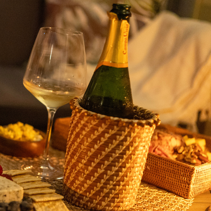 A beautifully handcrafted Julia Pine Needle Basket holding a chilled bottle of champagne, surrounded by a cozy charcuterie setup with a glass of white wine and soft lighting.