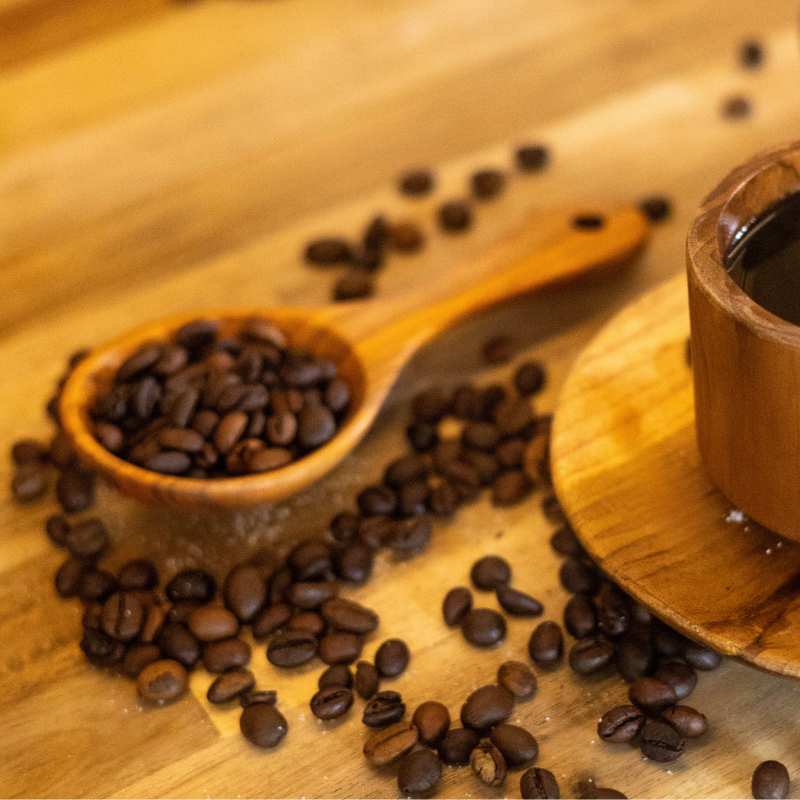 Handcrafted hardwood coffee scoop filled with roasted coffee beans, placed next to a wooden cup of coffee on a natural wooden surface, surrounded by scattered coffee beans for a warm, rustic ambiance.