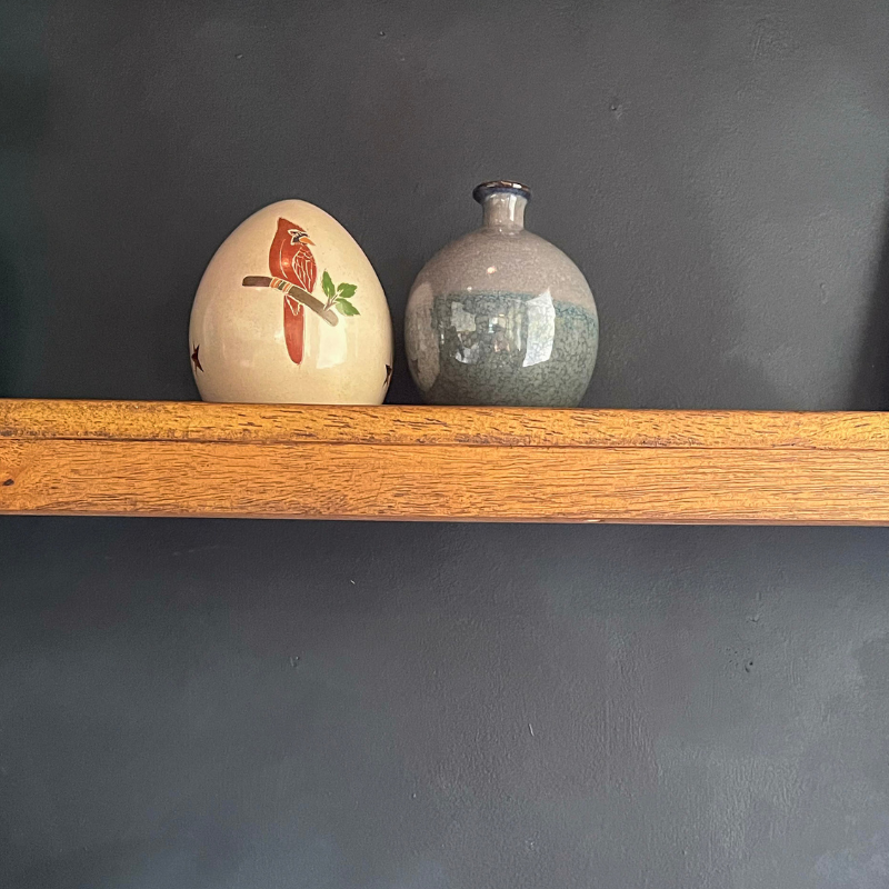 Spring Cardinal Luminary displayed on a wooden shelf, paired with a ceramic vase, showcasing its handcrafted design and vibrant cardinal artwork against a dark gray wall.