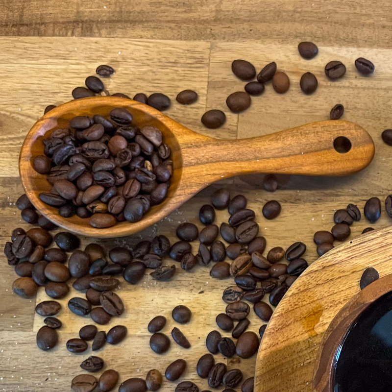 Handcrafted hardwood coffee scoop filled with dark roasted coffee beans, surrounded by scattered beans on a natural wooden surface, showcasing its rustic and practical design.