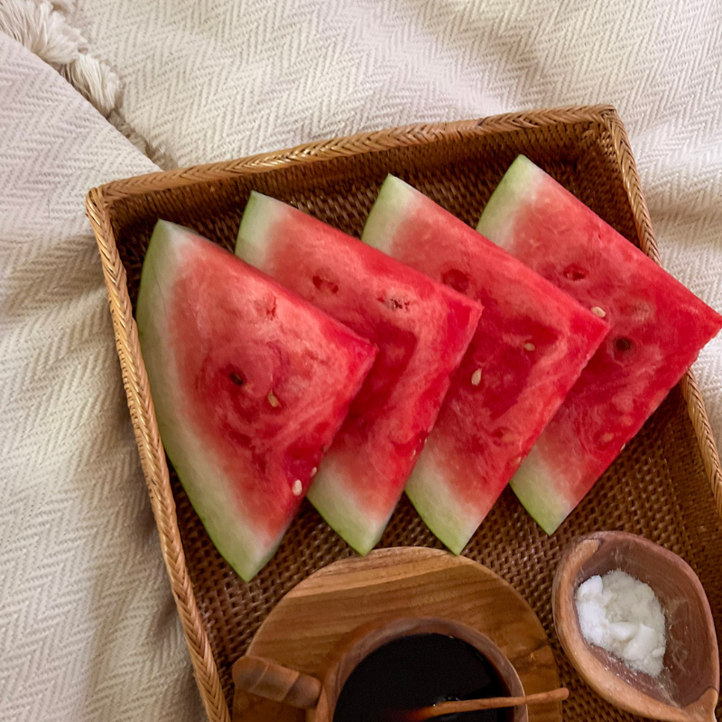 Handwoven rattan serving tray holding watermelon slices, a small wooden bowl of salt, and a teak coffee cup, placed on a soft cream blanket for a cozy presentation.