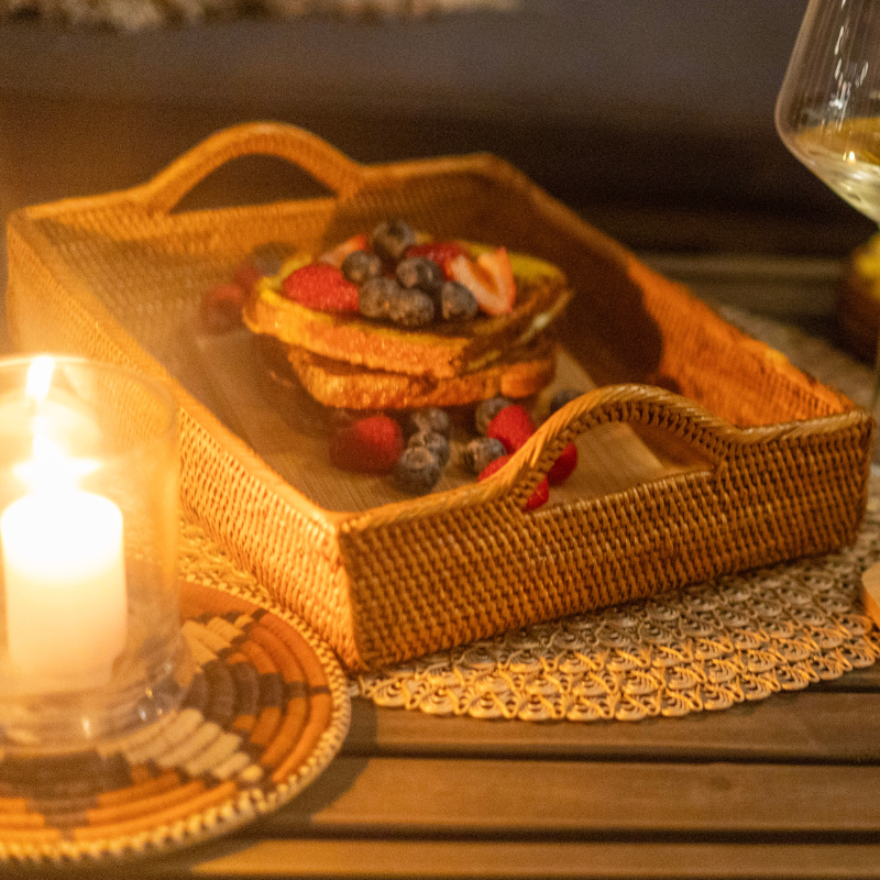 Handwoven rattan serving tray with handles, styled as a breakfast tray with French toast topped with fresh berries, placed on a woven mat beside a glowing candle for an inviting ambiance.