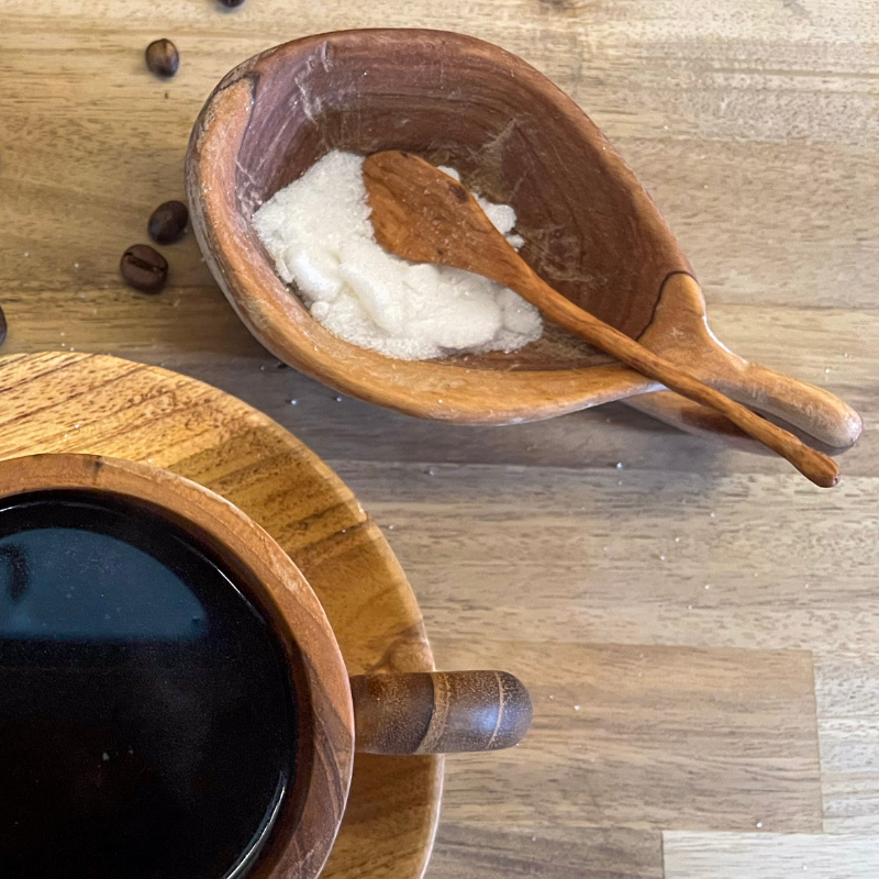 Handcrafted wooden spice bowl with a matching spoon, filled with sugar and placed on a rustic wooden surface alongside a wooden coffee cup, creating a warm and natural kitchen scene.