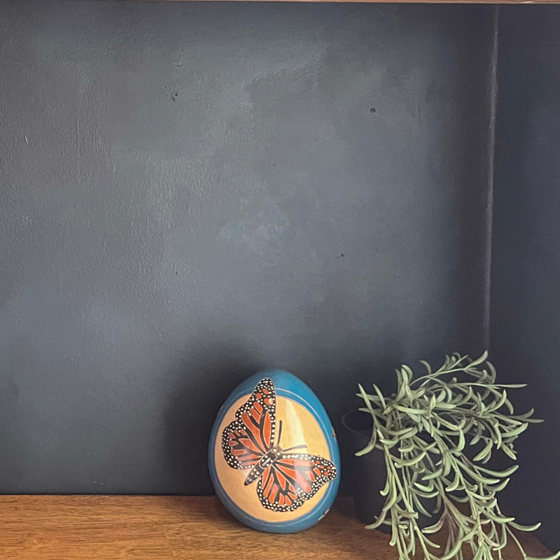 Handcrafted ceramic luminary with a vibrant butterfly design, displayed on a wooden surface against a dark gray background, paired with a green plant for a natural and elegant decor touch.